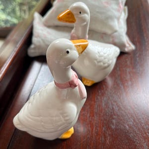 May include: Two white ceramic goose figurines with yellow beaks and feet. The goose on the left has a pink ribbon around its neck. The geese are sitting on a wooden surface.
