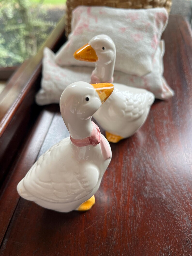 May include: Two white ceramic goose figurines with yellow beaks and feet. The goose on the left has a pink ribbon around its neck. The geese are sitting on a wooden surface.