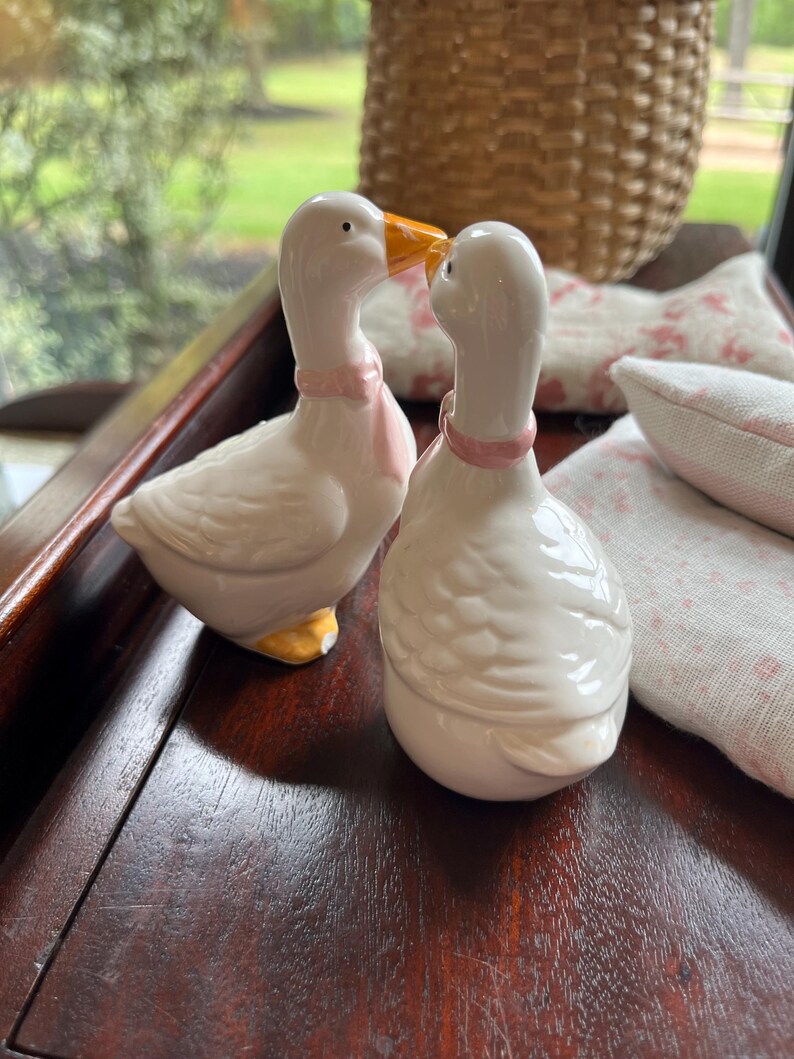 May include: A pair of white ceramic goose salt and pepper shakers with pink bows and orange beaks. The geese are facing each other and appear to be kissing.