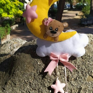 Può includere: Una luna crescente di feltro giallo con un fiocco di feltro rosa e un orso di feltro marrone al suo interno. La luna è appesa a uno spago bianco ed è attaccata a una nuvola di feltro bianco con un fiocco di feltro rosa. Tre stelle di feltro rosa sono appese al fiocco.