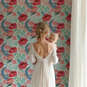 May include: A woman in a white dress holds a baby in front of a wall with a floral wallpaper pattern. The wallpaper features red, pink, and blue flowers with green leaves on a white background.