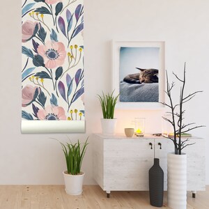 Op de afbeelding: Een witte muur met een rol bloemenbehang, een witte commode met een zwarte vaas en een witte vaas, een plant in een pot en een ingelijst beeld van een slapende kat.