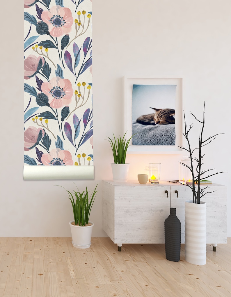 Op de afbeelding: Een witte muur met een rol bloemenbehang, een witte commode met een zwarte vaas en een witte vaas, een plant in een pot en een ingelijst beeld van een slapende kat.