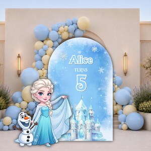 Op de afbeelding: Een boogvormige achtergrond met een blauw aquarelontwerp met witte sneeuwvlokken, een kasteelillustratie en de tekst Alice turns 5. Een tekenfilmfiguur met blond haar in een blauwe jurk en een sneeuwpop staan ervoor.