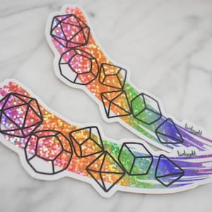 May include: Two die-cut stickers featuring a rainbow-colored, glittery brushstroke background with black line art illustrations of various polyhedral dice. Each sticker includes the small text "livelygold" printed near the bottom right edge.