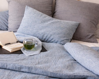May include: A bed with light blue linen bedding, including a duvet, pillows, and a rolled-up blanket. A clear glass cup of tea and an open notebook rest on the bed. The pillows are various shades of gray and blue.