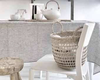 Könnte beinhalten: Ein neutrales Interieur mit einer weißen Teekanne mit Holzgriff, einem geflochtenen Korb und einer gestreiften Tischdecke. Regale darüber zeigen Bücher und dekorative Gegenstände, die eine minimalistische Ästhetik schaffen.