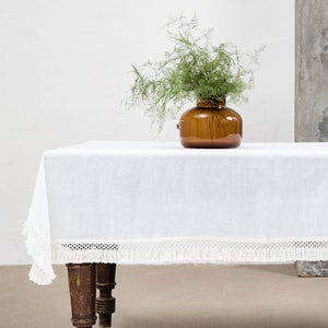 May include: A white tablecloth with a lace trim and fringe hangs over a wooden table with turned legs. A vase with green foliage sits on the table. A small wooden stool with a round top sits in the background.