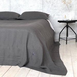 May include: A dark charcoal grey bedspread covers a bed with two matching pillows. The fabric has a visible weave texture and a small rectangular tag near the bottom corner. The bed is positioned next to a black metal side table with three legs.