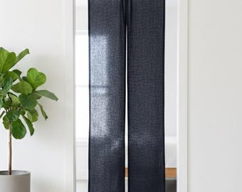 Puede incluir: Una cortina de puerta de tela gris carbón oscuro con un tejido texturizado, que cuelga verticalmente de una barra negra dentro de un marco de puerta blanco. La cortina está dividida en dos paneles, permitiendo que la luz pase a través del material.