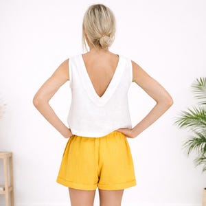 May include: A sleeveless white top with a deep V-neck paired with yellow shorts. The shorts have a relaxed fit and a rolled hem. The outfit is simple and summery, perfect for warm weather.