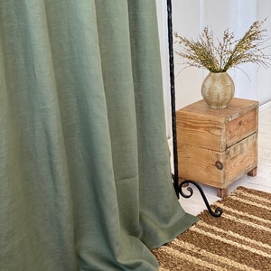 Pode incluir: Uma cortina de linho verde-s&aacute;lvia pendurada na frente de uma mesa auxiliar de madeira com um vaso de flores secas. A cortina est&aacute; reunida na parte inferior e a mesa &eacute; feita de madeira recuperada.