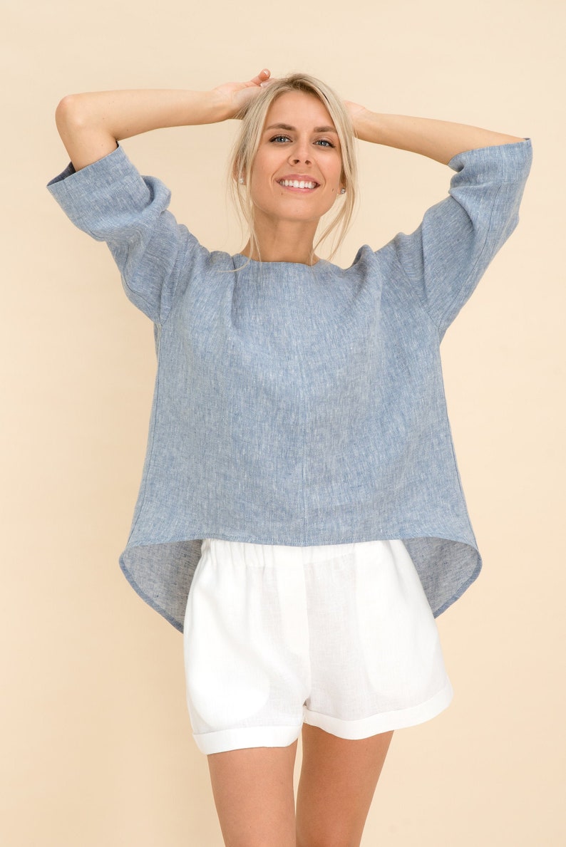 May include: A light blue linen top with three-quarter sleeves and a rounded hemline, paired with white linen shorts. The top has a relaxed fit, and the shorts have an elastic waistband. The outfit is casual and summery.
