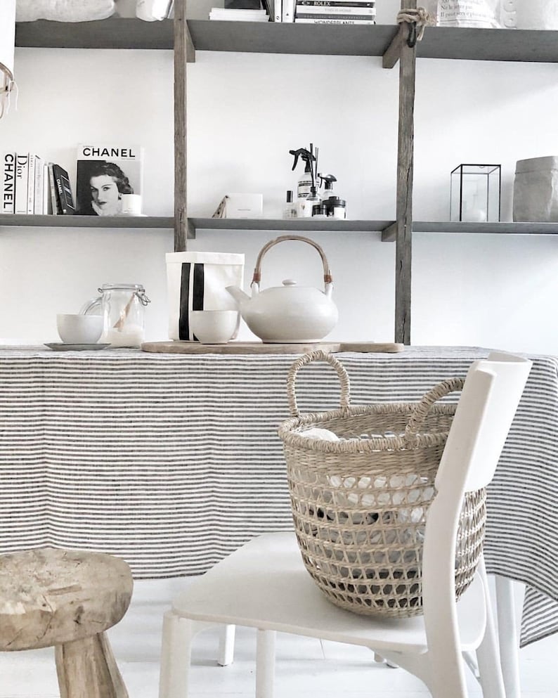 May include: A neutral-toned interior scene featuring a white teapot with a wooden handle, a woven basket, and a striped tablecloth. The shelves above display books and decorative items, creating a minimalist aesthetic.