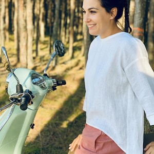 May include: A light green vintage scooter with a chrome mirror and black handlebars. The scooter is parked in a wooded area. A person is wearing a white long-sleeve shirt and pink shorts.