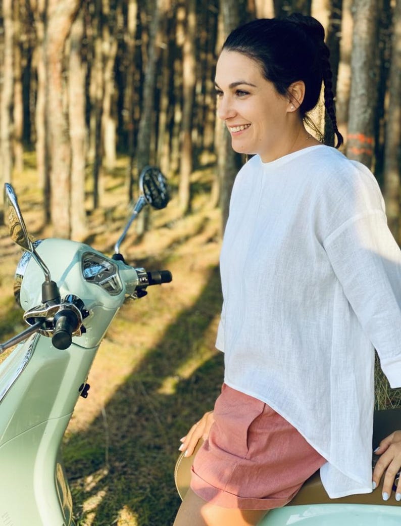 May include: A light green vintage scooter with a chrome mirror and black handlebars. The scooter is parked in a wooded area. A person is wearing a white long-sleeve shirt and pink shorts.