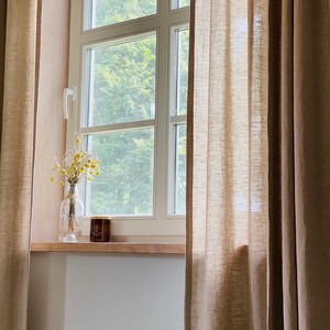 K&ouml;nnte beinhalten: Ein Paar bodenlange, beige Leinenvorh&auml;nge rahmen ein wei&szlig;es Fenster mit Holzsims ein. Auf dem Sims steht eine durchsichtige Glasflasche mit getrockneten gelben Blumen und ein kleines braunes Kerzenglas mit einem Holzdeckel.