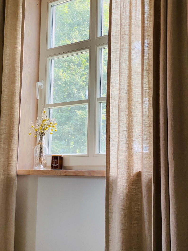 K&ouml;nnte beinhalten: Ein Paar bodenlange, beige Leinenvorh&auml;nge rahmen ein wei&szlig;es Fenster mit Holzsims ein. Auf dem Sims steht eine durchsichtige Glasflasche mit getrockneten gelben Blumen und ein kleines braunes Kerzenglas mit einem Holzdeckel.