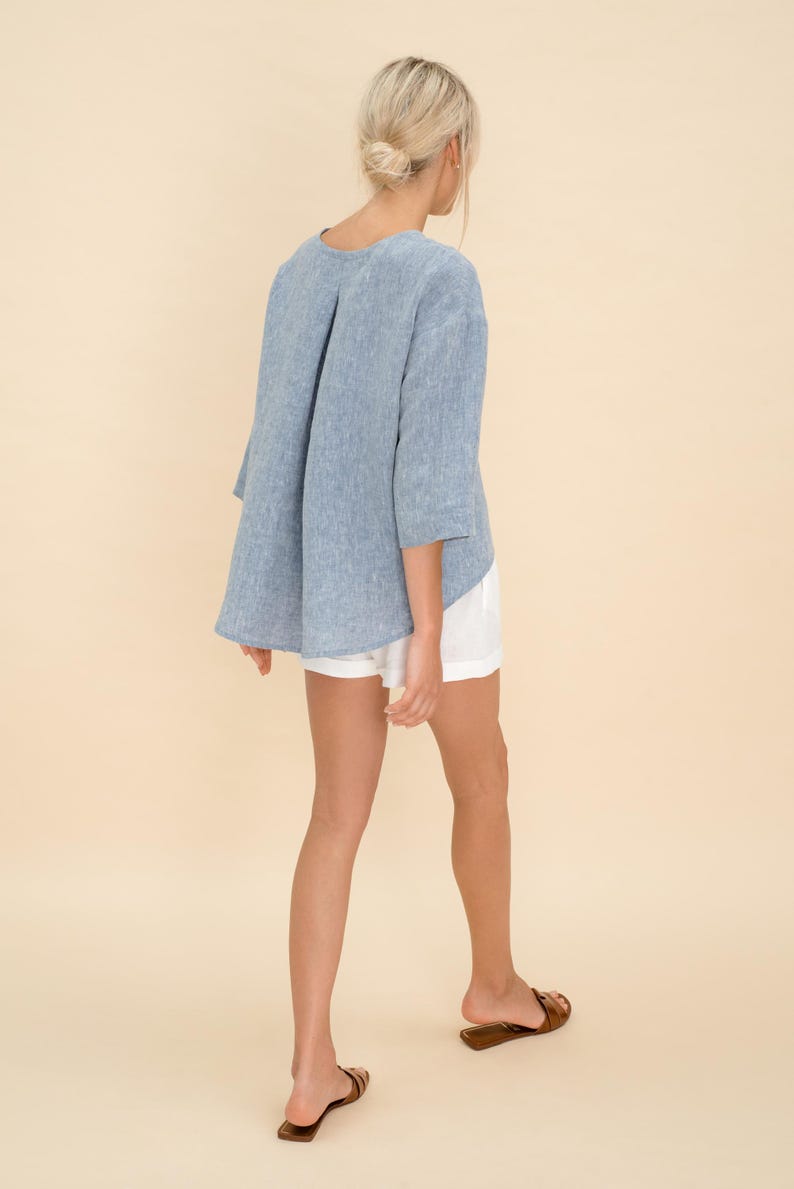 May include: A light blue linen top with three-quarter sleeves and a pleated back detail. The top is paired with white shorts and brown sandals. The outfit is casual and suitable for warm weather.