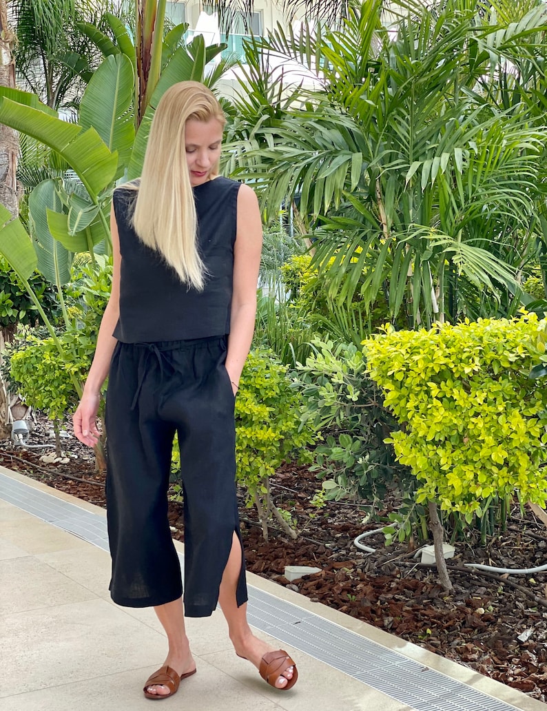 May include: A black linen outfit consisting of a sleeveless top and cropped pants with side slits. The pants have a drawstring waist. The outfit is paired with brown leather sandals. The background features lush green foliage and a building.