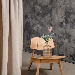 K&ouml;nnte beinhalten: Ein heller Holzstuhl mit geteilter R&uuml;ckenlehne und Sitzfl&auml;che, auf dem eine durchsichtige Glaskaraffe steht. Die Karaffe enth&auml;lt einen einzelnen St&auml;ngel mit kleinen wei&szlig;-rosa Bl&uuml;ten und gr&uuml;nen Bl&auml;ttern und ist mittig auf dem Sitz platziert.