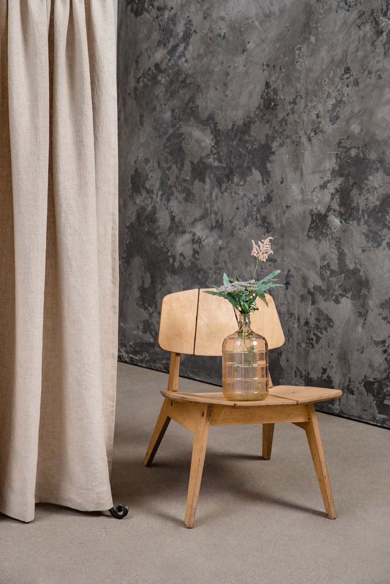 K&ouml;nnte beinhalten: Ein heller Holzstuhl mit geteilter R&uuml;ckenlehne und Sitzfl&auml;che, auf dem eine durchsichtige Glaskaraffe steht. Die Karaffe enth&auml;lt einen einzelnen St&auml;ngel mit kleinen wei&szlig;-rosa Bl&uuml;ten und gr&uuml;nen Bl&auml;ttern und ist mittig auf dem Sitz platziert.
