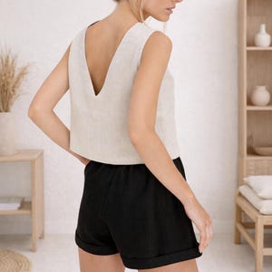 May include: A sleeveless, beige linen top with a deep V-neck paired with black linen shorts. The shorts have a rolled hem. The outfit is casual and comfortable, suitable for warm weather. The model is wearing the outfit in a light-filled room.