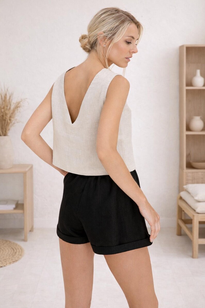 May include: A sleeveless, beige linen top with a deep V-neck paired with black linen shorts. The shorts have a rolled hem. The outfit is casual and comfortable, suitable for warm weather. The model is wearing the outfit in a light-filled room.