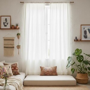 K&ouml;nnte beinhalten: Ein Paar halbtransparente wei&szlig;e Vorh&auml;nge, die an einer Holzstange &uuml;ber einem Fenster h&auml;ngen. Die Vorh&auml;nge sind lichtdurchl&auml;ssig und lassen sanftes Licht hindurch. Sie befinden sich &uuml;ber einem flachen beigen Bodenkissen und einem Jute-Teppich.