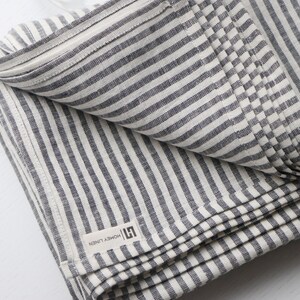 May include: Folded linen textile with alternating gray and white stripes. The fabric has a textured appearance and a label that reads "HONEY LINEN". The textile is folded neatly, showcasing the striped pattern.