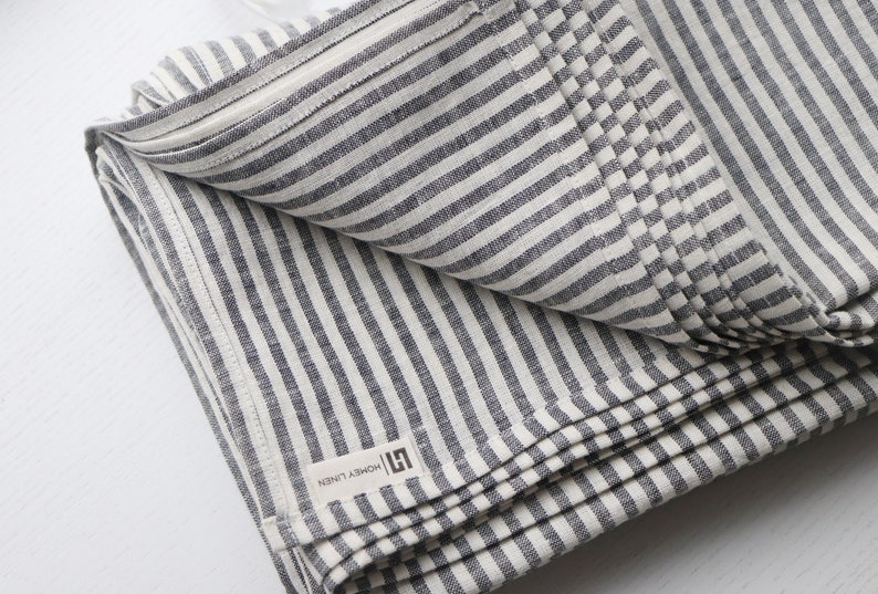May include: Folded linen textile with alternating gray and white stripes. The fabric has a textured appearance and a label that reads "HONEY LINEN". The textile is folded neatly, showcasing the striped pattern.