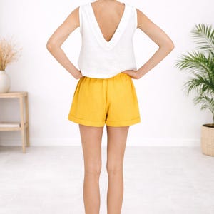 May include: A sleeveless white top with a deep V-neck paired with yellow shorts. The shorts have a relaxed fit and a rolled hem. The outfit is simple and summery, perfect for warm weather.