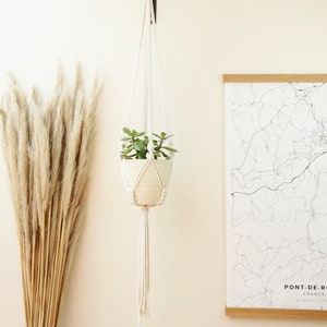 May include: A white macrame plant hanger with a small potted plant hanging from it. The hanger is attached to a hook on a white wall. There is a dried pampas grass bouquet to the left of the hanger and a framed map of Pont-de-Roide, France to the right.