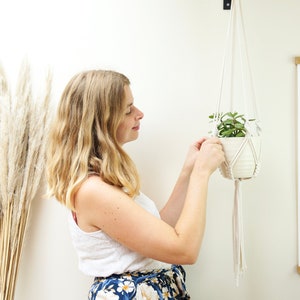 May include: A woman hangs a macrame plant hanger with a white ceramic pot and a small green plant inside. The hanger is made of natural white cotton cord. The plant hanger is hanging from a hook on a white wall.