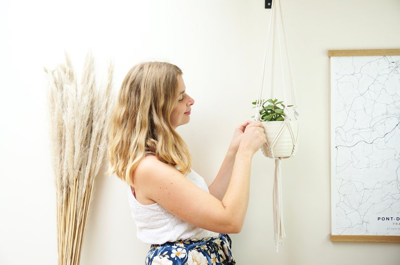 May include: A woman hangs a macrame plant hanger with a white ceramic pot and a small green plant inside. The hanger is made of natural white cotton cord. The plant hanger is hanging from a hook on a white wall.