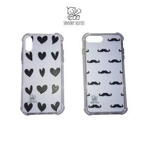 May include: Two clear phone cases with black designs. One case has a pattern of black hearts on a white background. The other case has a pattern of black mustaches on a white background. Both cases have the text "Sensory Selfies" printed on the bottom.