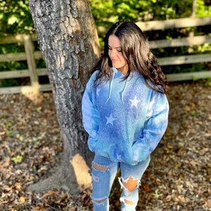 May include: A blue tie-dye hoodie with white stars printed on it. The hoodie has a drawstring hood and a kangaroo pocket. The model is wearing ripped jeans and white sneakers.