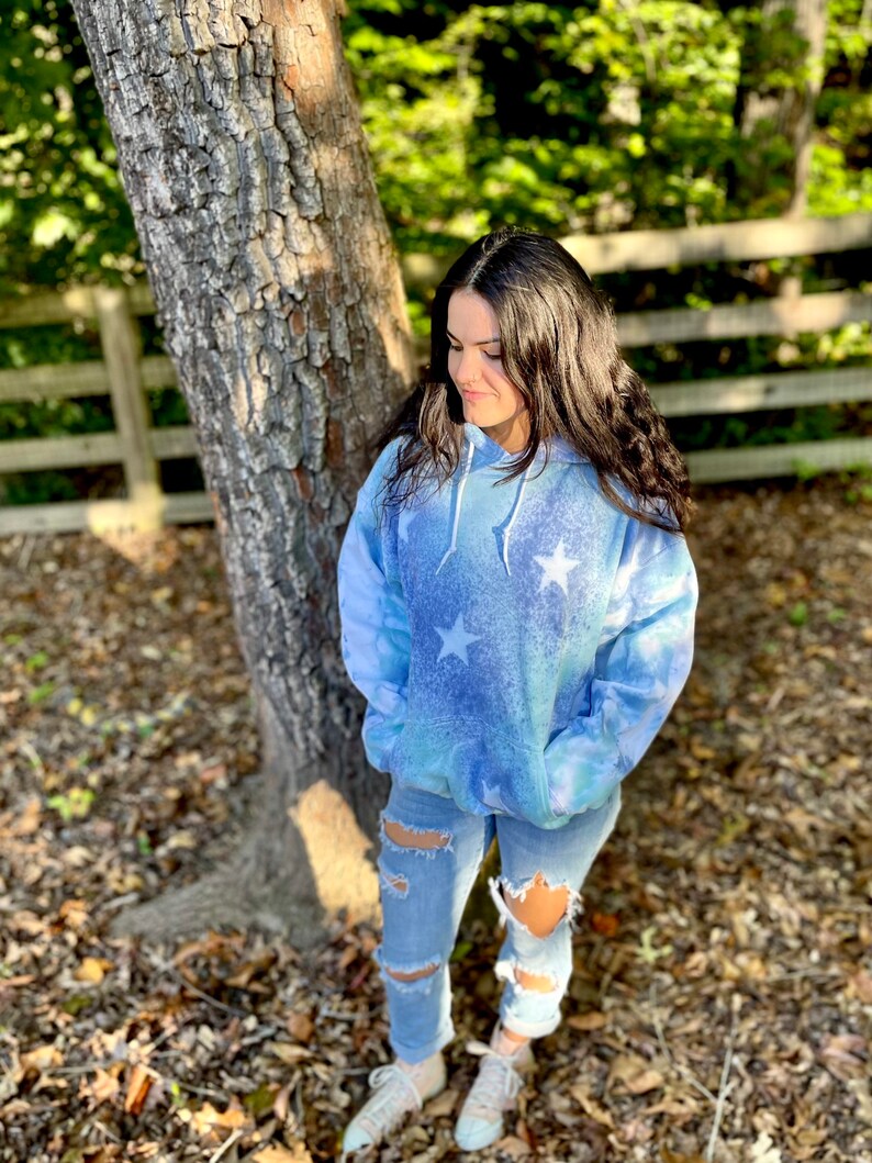 May include: A blue tie-dye hoodie with white stars printed on it. The hoodie has a drawstring hood and a kangaroo pocket. The model is wearing ripped jeans and white sneakers.