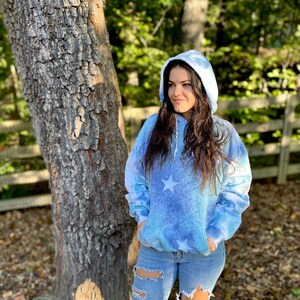 May include: A blue tie-dye hoodie with white stars. The hoodie has a drawstring hood and a kangaroo pocket. The model is wearing the hoodie with ripped jeans.