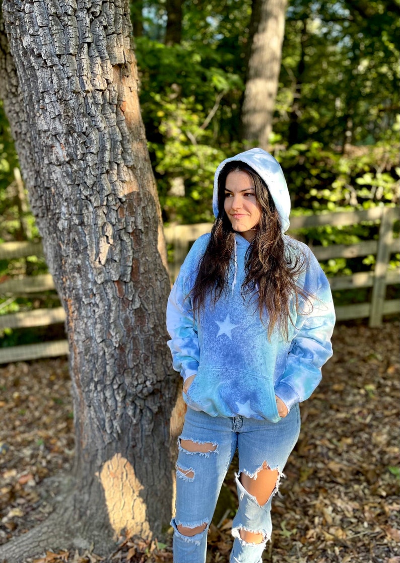 May include: A blue tie-dye hoodie with white stars. The hoodie has a drawstring hood and a kangaroo pocket. The model is wearing the hoodie with ripped jeans.