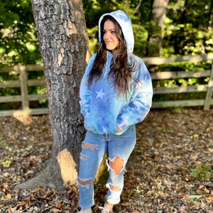 May include: A blue tie-dye hoodie with a white star design. The hoodie has a drawstring hood and a kangaroo pocket. The model is wearing the hoodie with ripped jeans and white sneakers.