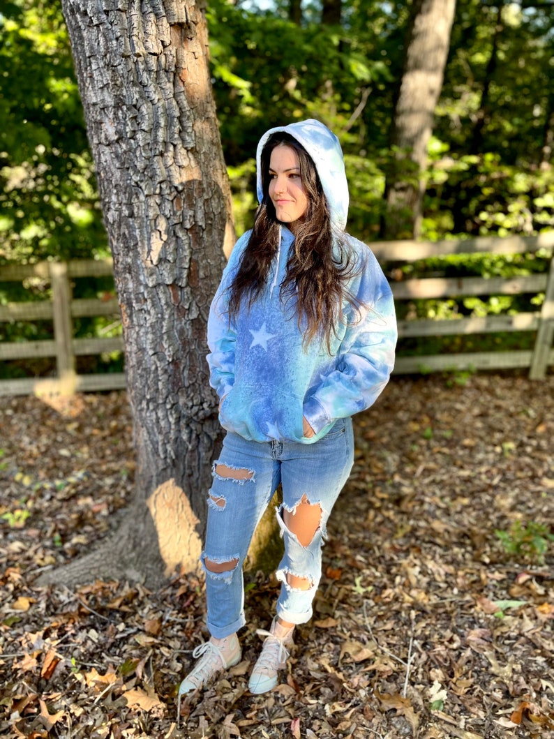 May include: A blue tie-dye hoodie with a white star design. The hoodie has a drawstring hood and a kangaroo pocket. The model is wearing the hoodie with ripped jeans and white sneakers.