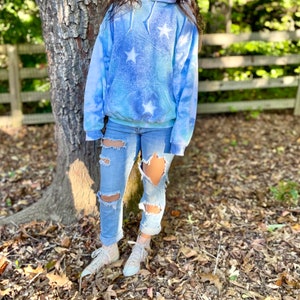 May include: A light blue tie-dye hoodie with white stars. The hoodie has a drawstring hood and a kangaroo pocket. The model is wearing ripped jeans and white sneakers.