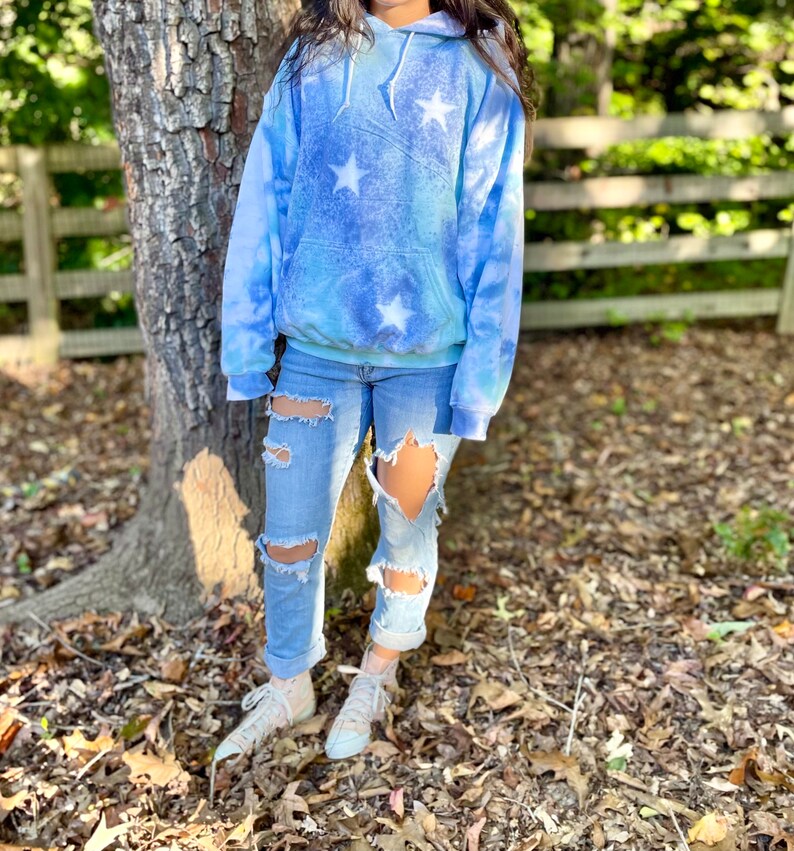 May include: A light blue tie-dye hoodie with white stars. The hoodie has a drawstring hood and a kangaroo pocket. The model is wearing ripped jeans and white sneakers.