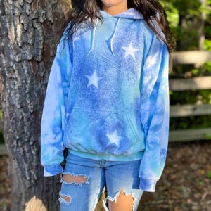 May include: A blue and white tie-dye hoodie with white stars. The hoodie has a drawstring hood and a kangaroo pocket.