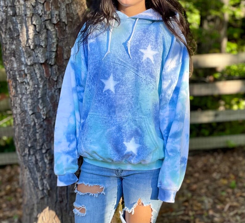 May include: A blue and white tie-dye hoodie with white stars. The hoodie has a drawstring hood and a kangaroo pocket.