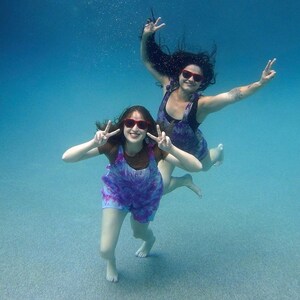 May include: Two people wearing tie-dye dresses and red sunglasses pose for a photo while underwater. They are both making peace signs with their hands.