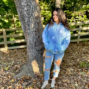 May include: A woman wearing a blue tie-dye hoodie with white stars and ripped denim jeans. She is standing in front of a tree with a wooden fence behind her.