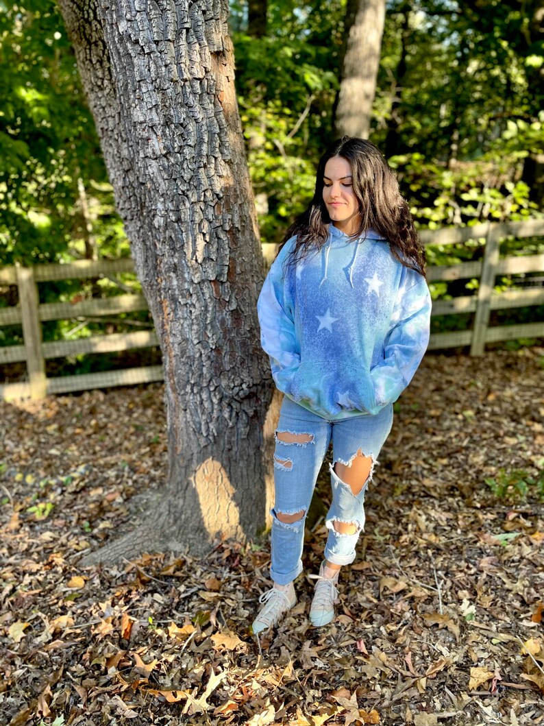 May include: A woman wearing a blue tie-dye hoodie with white stars and ripped denim jeans. She is standing in front of a tree with a wooden fence behind her.