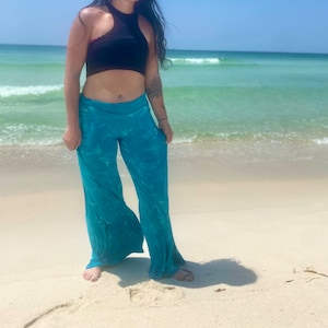 May include: A woman wearing a black halter top and blue tie-dye wide-leg pants stands on a white sandy beach with turquoise water in the background.